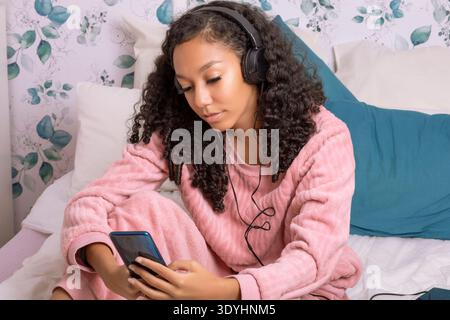 Eine junge Frau sitzt bequem in ihrem gemütlichen Schlafzimmer, trägt einen rosafarbenen Mantel und Kopfhörer. Sie konzentriert sich auf ihr Smartphone und genießt ihre Lieblingsmusik Stockfoto