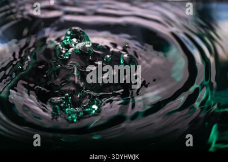 372.Nahaufnahme der dunklen Wasseroberfläche mit aufsteigender zentraler Blase und konzentrischen Wellen. Grüne und türkisfarbene Reflexe schaffen abstrakte Textur. Leicht Stockfoto