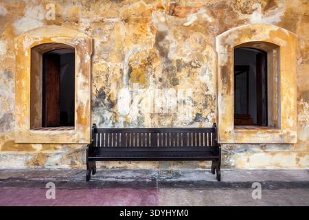 Eine einfache Holzbank sitzt zwischen zwei Bogenfenstern vor einer strukturierten, verwitterten Wand im Castillo San Cristobal in Old San Juan, Puerto Rico. T Stockfoto