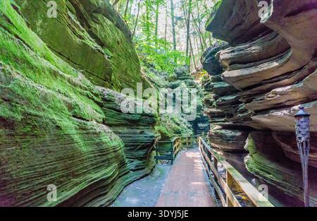 Ein schmaler Pfad schlängelt sich durch einen felsigen Canyon. Der Weg ist gesäumt von Bäumen und Moos, und es gibt eine hölzerne Brücke, die einen kleinen Bach überquert. Die Szene Stockfoto