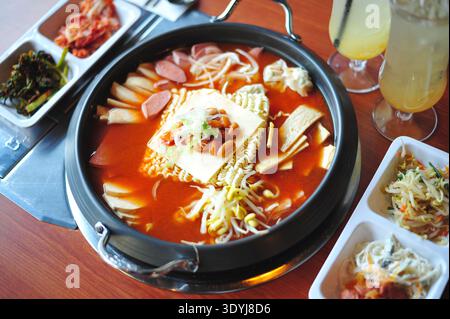 Traditioneller koreanischer Eintopf der Armee (Budae Jjigae) mit Ramen-Nudeln, Wurst, Schinken, Tofu und Käse in scharfer Brühe serviert mit koreanischen Beilagen, ein beliebtes Stockfoto