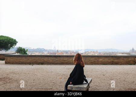 Junge Frau, die auf einer Bank sitzt und die Skyline der Stadt von der Aussichtsterrasse in Rom betrachtet. Reise- und Kontemplationskonzept. Stockfoto
