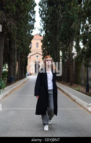 Stilvolle junge Frau, die in einer ruhigen Parkallee mit hohen Bäumen und historischer Architektur im Hintergrund schlendert. Stockfoto