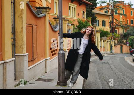 Junge rothaarige Frau, die sich an einen Straßenmast in einem bunten europäischen Viertel lehnt. Urbanes Lifestyle-Konzept. Stockfoto
