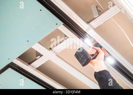 Nahaufnahme einer Hand mit einem Akku-Bohrer zur Installation eines abgehängten Deckenrahmens mit LED-Schienenbeleuchtung in einem modernen Innenraum. Stockfoto