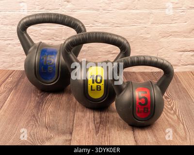 Drei vinylbeschichtete Wasserkettlebells in Blau Gelb und Rot auf Holzboden vor einer weißen Ziegelwand für Heimtraining im Fitnessstudio. Stockfoto