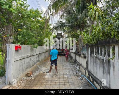 Ein Arbeiter gießt Beton auf einen neu angelegten Weg, der von üppiger Vegetation in Bang Lamung umgeben ist. Die tropische Atmosphäre zeigt Palmen und farbenfrohe Pflanzen unter einem strahlenden Himmel. Stockfoto