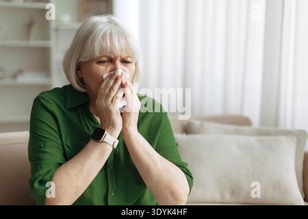 Ältere Frau, die zu Hause auf dem Sofa sitzt, mit Taschentuch in die Nase bläst Stockfoto