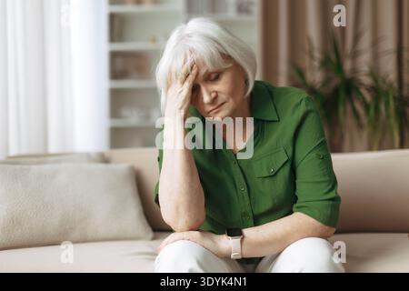 Ältere Frau, die auf dem Sofa sitzt, mit besorgtem Ausdruck und Kopf hält Stockfoto