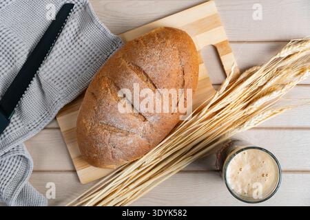 Runder rustikaler Brotlaib mit Mehlkruste neben einem Glas Kwass, ergänzt durch Weizenstiele auf Holzoberfläche für eine gesunde Szene Stockfoto