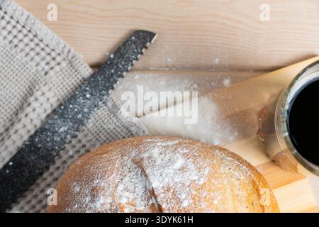 Runder rustikaler Brotlaib mit Mehlkruste neben einem Glas Kwass, ergänzt durch Weizenstiele auf Holzoberfläche für eine gesunde Szene Stockfoto
