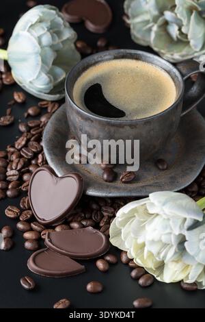 Graubraune Grunge Tasse frischer heißer Kaffee mit Bohnen, Frottee Tulpenblüten und dunkle Schokoladenstücke auf schwarzem Hintergrund. Schokoladenstück in Form hören Stockfoto