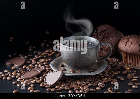 Graubraune Grunge Tasse frischer heißer Kaffee mit Dampf und Bohnen, dunkle Schokoladenstücke, Muffins auf schwarzem Hintergrund. Schokoladenstück in Form Herz und Stockfoto