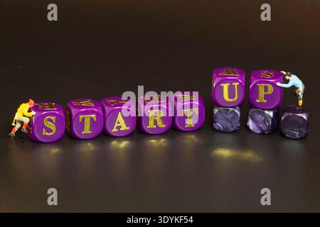 Miniaturkletterer drücken lila Buchstabenwürfel und bilden das Wort START-UP in einer Studio-Makroszene, die Unternehmertum und Neuanfänge symbolisiert. Stockfoto