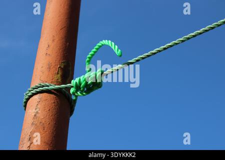 Ein grünes Seil ist an einen rostigen Metallstab gebunden. Das Seil ist mit einem Knoten gesichert. Die Sonne scheint und der Himmel ist klar, ohne Wolken zu sehen. Stockfoto