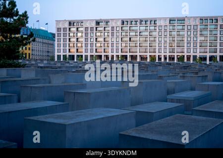 Graue Betonstelen der Gedenkstätte für die ermordeten Juden Europas füllen die Szene, ein modernes Gebäude erhebt sich dahinter, Abendfenster leuchten, Berlin, breit Stockfoto