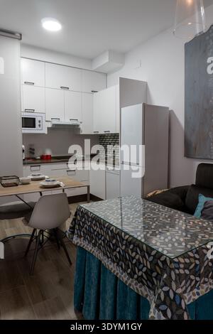 Saubere, moderne Küche und Essecke mit weißen Schränken, Geräten, Esstisch und Möbeln im Wohnbereich Stockfoto
