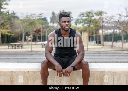 Muskulöser afroamerikanischer männlicher Athlet in schwarzer Activewear, der an der Grenze in der Stadt sitzt und nach intensivem Fitnesstraining ausruht Stockfoto