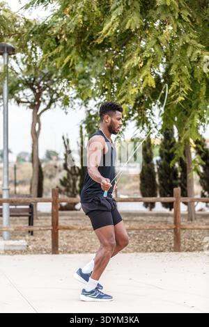 Seitenansicht des gesamten Körpers eines jungen afroamerikanischen Athleten beim Sport-Outfit-Training mit Seilspringen in der Nähe von grünen Bäumen während des Fitnesstrainings Stockfoto