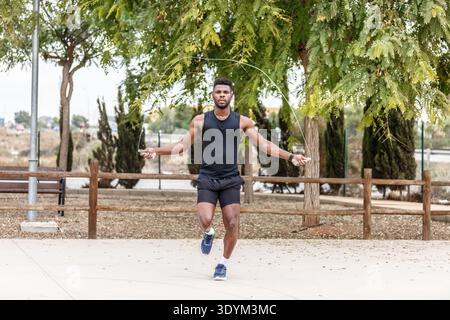 Ganzkörper eines bärtigen afroamerikanischen Sportlers in schwarzer Aktivkleidung, der während des Fitnesstrainings im Sommerpark mit einem Seil gegen grüne Nadelbäume springt Stockfoto