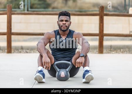 Voller Körper eines jungen bärtigen muskulösen afroamerikanischen Athleten in Sportbekleidung, der mit einer Kettlebell auf dem Boden sitzt und in die Kamera blickt Stockfoto