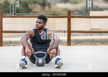 Voller Körper eines entschlossenen ethnischen Athleten in Sportbekleidung und Turnschuhen, der während des Fitness-Trainings im Freien mit einer Kettlebell auf dem Boden sitzt und wegblickt Stockfoto