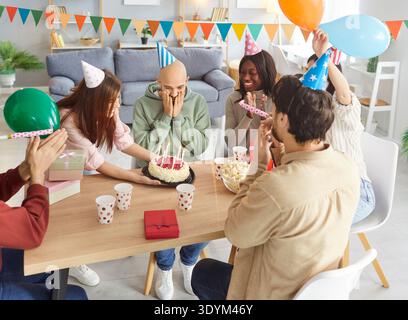 Aufgeregter Mann, der sein Gesicht mit Händen bedeckt, während Freunde in Partyhüten ihn mit Geburtstagskuchen überraschen. Stockfoto