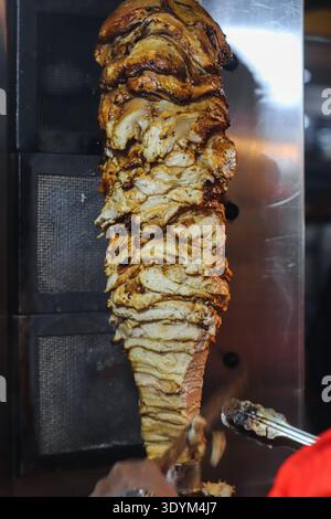 Vertikale Nahaufnahme des Händlers, der geröstete Hühnchen-Shawarma-Kebab aus rotierendem Spieß schneidet, mit saftigen Schichten und authentischer Street Cuisine-Zubereitung. Stockfoto