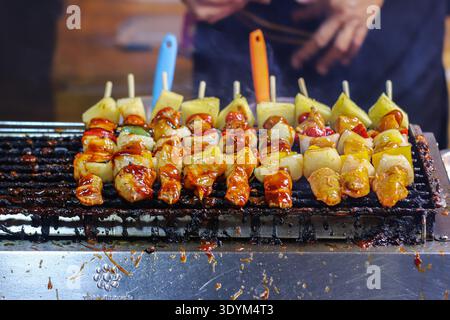 Thailändische Grillspieße mit Hähnchen Ananaszwiebeln und Paprika, die über Holzkohle gegrillt werden, an einem belebten Nachtmarkt Street Cuisine Stand. Stockfoto