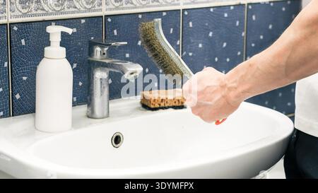 Nahaufnahme einer muskulösen Hand, die eine Messingdrahtbürste über einem weißen Waschbecken mit Chromarmatur, Seifenspender und Schwamm in einem gefliesten Badezimmer hält. Stockfoto