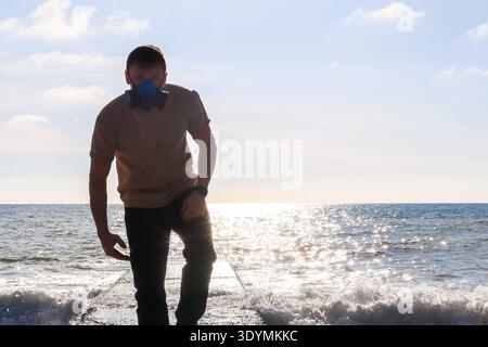Individuelle Gesichtsmaske spaziert lässig auf dem Pier am Meer, umgeben von schäumenden Wellen und Horizontlicht Stockfoto