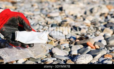 Detaillierte Ansicht einer Hand mit Handschuhen, die Kunststoffabfälle von einer felsigen Strandoberfläche hebt. Stockfoto