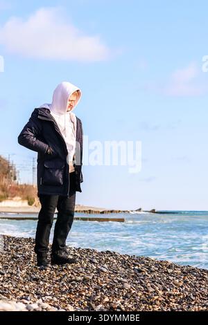 Individual in Kapuzenoberbekleidung blickt auf den Kiesstrand mit Meereswellen in der Nähe. Stockfoto