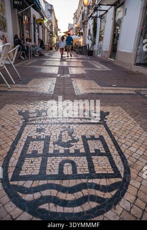 Eine Straße in Faro, Portugal, mit einem dekorativen Mosaik im Vordergrund, das das Stadtwappen darstellt. Faro ist die Hauptstadt der Algarve in Stockfoto