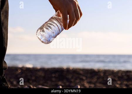 Hand in legere Kleidung, die Plastikflasche am Hals gegen den ruhigen Meereshorizont und das felsige Ufer anschließt. Stockfoto