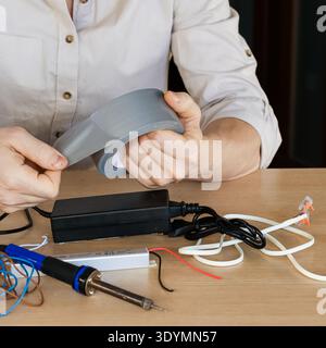 Aus nächster Nähe sehen Sie, wie die Hände einen Streifen grauen Klebebandes über einem Tisch mit Lötwerkzeugen, Drähten und Stromversorgungseinheiten in einer Bastelszene ziehen und reißen. Stockfoto