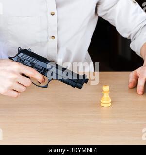 Nahaufnahme der Hand einer Person, die eine schwarze Pistole packt, die direkt auf einen kleinen gelben Schachpfand auf Holzoberfläche zeigt Stockfoto