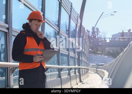 Fokussierter Bauprofi in Schutzhelm und Weste inspiziert Dokumente in der Nähe von reflektierenden Glaswänden auf urbaner Infrastruktur im Winter. Stockfoto