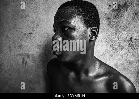 Schwarz-weiße Nahaufnahme-Porträt eines jungen afrikanischen Tänzers, der während einer erholsamen Pause mit Schweiß bedeckt ist und einen Moment ruhiger Intensität reflektiert. Stockfoto
