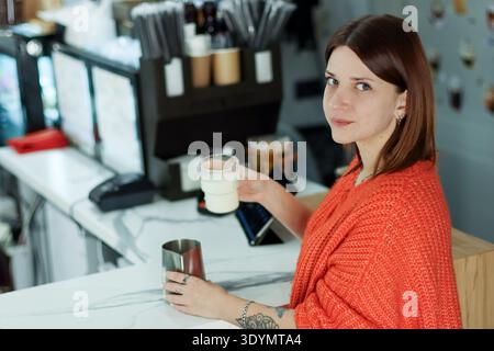 Der Barista mit tätowiertem Arm in einem gemütlichen orangefarbenen Pullover gießt weiße Milchflüssigkeit aus der Tasse in den Krug auf die Kaffeekasse aus weißem Marmor. Stockfoto