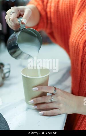 Nahaufnahme von tätowierten Händen in orangefarbenen Ärmeln, die cremige Milchschaummilch in eine Kaffeetasse aus Papier auf der Marmortheke gießen. Stockfoto