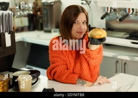 Der Barista in einem gemütlichen orangefarbenen Pullover hält frisch gebackenes Croissant neben Tassen und Ausrüstung auf weißem Marmor. Stockfoto