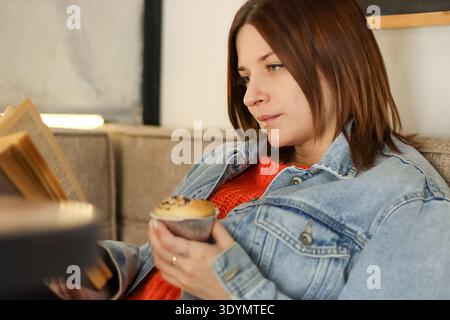 Profilansicht einer jungen Frau in Jeansjacke, die Buch- und Schokoladenmuffin während der Kaffeepause genießt Stockfoto