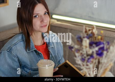 Junge Frau in lässiger Kleidung, die entspannt im Café sitzt, Eiskaffee zum Mitnehmen hält und ein Buch mit getrockneten Blumen im Vordergrund Stockfoto