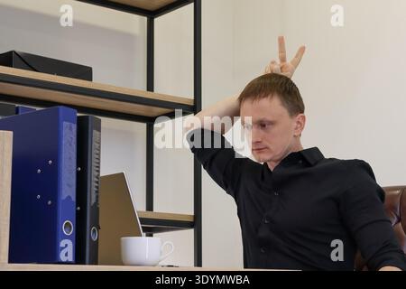 Sitzender junger Profi im Büro formt Hörner mit Fingern auf dem Kopf und blickt ernsthaft in die Kamera. Stockfoto