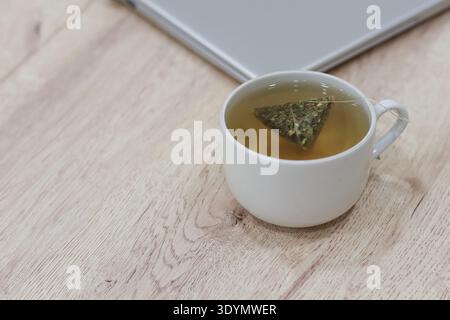Nahaufnahme von grünem Kräutertee, der in heißem Wasser in einem Keramikbecher neben dem Laptop auf hellem Holz infundiert wird, Büroeinrichtung Stockfoto
