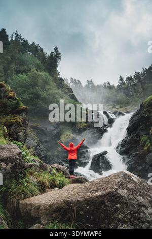 Fröhliche Trekkerin in leuchtend roter wasserdichter Jacke, die den spektakulären Låtefossen-Wasserfall in Norwegen genießt und auf einer Klippe mit fließenden cas steht Stockfoto