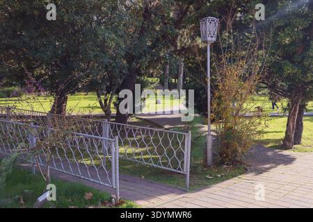 Elegantes weißes schmiedeeisernes Tor mit geschwungenen Motiven führt in einen üppig grünen Park mit hohen Bäumen, Sträuchern und dekorativen Laternen. Stockfoto