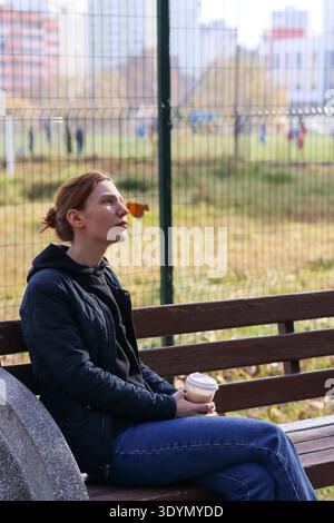 Profil einer Frau in schwarzer Jacke und blauer Jeans, die im Park sitzt, einen Drink hält und in der herbstlichen Landschaft in den Himmel blickt Stockfoto