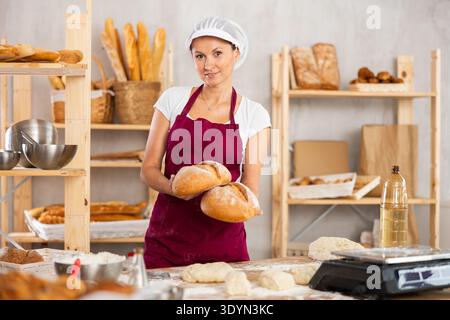 Junge Bäckerinnen stehen in der Nähe des Sortiments in der Bäckereiküche mit Korbbrot Stockfoto